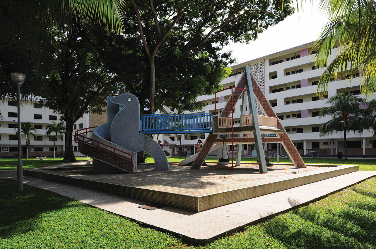 Outdoor playground with slides and swings in front of apartment buildings and trees on a sunny day.